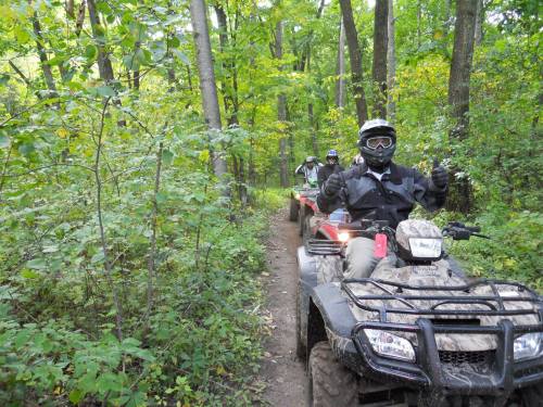 Two people riding ATVs on a narrow trail through a lush green forest, with one giving a thumbs-up.