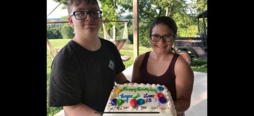 Two young people hold a birthday cake decorated with colorful icing, smiling outdoors in a sunny setting.