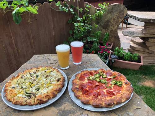 Two pizzas on a stone table: one with mushrooms and green peppers, the other with pepperoni and jalapeños, alongside two beers.