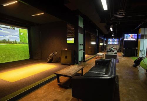 Indoor golf simulation area with multiple screens, seating, and golf equipment. Modern, spacious, and well-lit environment.