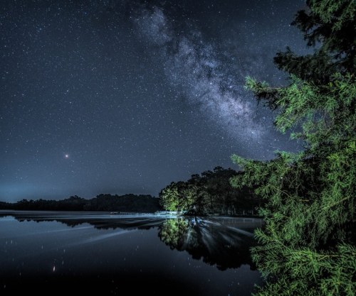 A serene night scene of a lake reflecting the Milky Way, framed by trees under a starry sky.