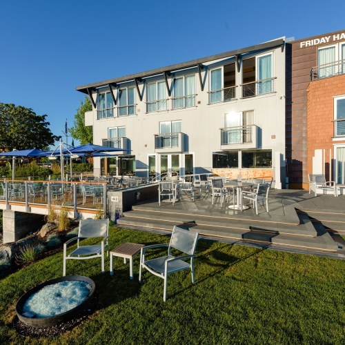A modern building with outdoor seating, surrounded by greenery and blue skies, near a waterfront area.