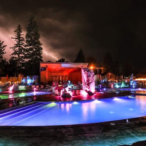 A vibrant pool area at night, illuminated with colorful lights, under a stormy sky with lightning in the background.