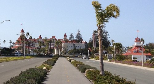 View of a palm-lined road leading to a historic hotel with red roofs and flags, surrounded by greenery and blue sky.
