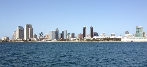 Panoramic view of a city skyline with modern buildings along a waterfront under a clear blue sky.