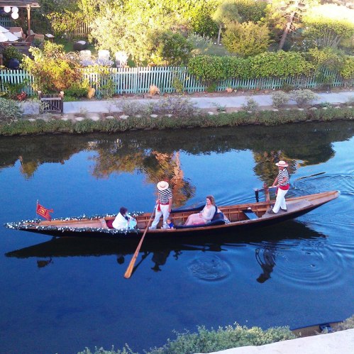 A gondola glides through a serene canal, with two rowers and a passenger enjoying the scenic surroundings.
