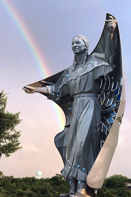 A large metallic statue of a woman with a rainbow in the background, showcasing intricate details and a flowing design.