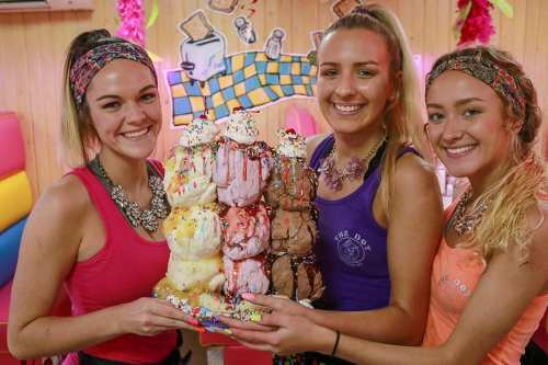 Three smiling women hold a large, colorful ice cream tower topped with sprinkles in a vibrant, playful setting.