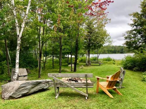 A serene outdoor scene with wooden benches, a fire pit, and trees by a calm lake under a cloudy sky.