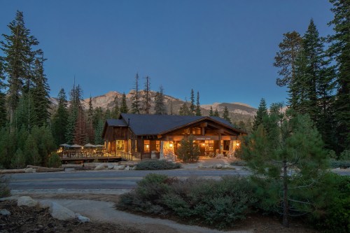A cozy mountain lodge illuminated at dusk, surrounded by tall trees and rocky peaks in the background.