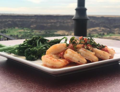 A plate of shrimp with tomatoes and herbs, served alongside sautéed broccolini, with a scenic view in the background.