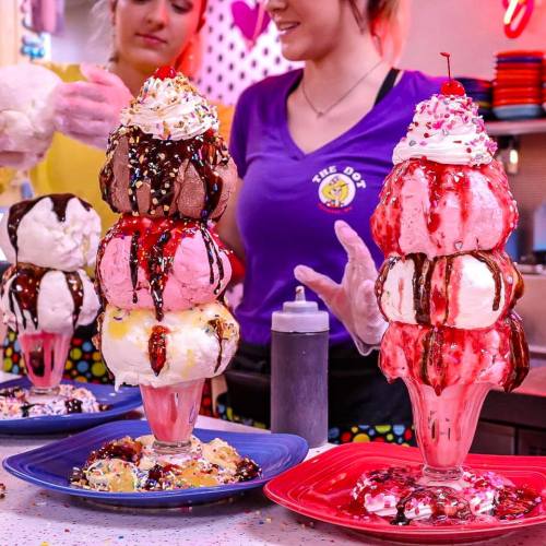 A towering ice cream sundae with multiple scoops, whipped cream, chocolate sauce, and a cherry on top, served in colorful dishes.