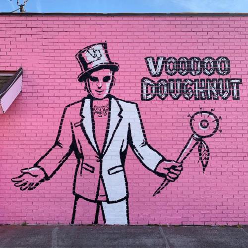 A vibrant pink mural featuring a stylized figure in a white suit and top hat, holding a wand, labeled "Voodoo Doughnut."