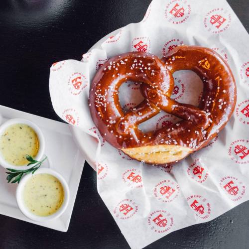 A large, soft pretzel sprinkled with salt, served with two dipping sauces on a branded paper and plate.