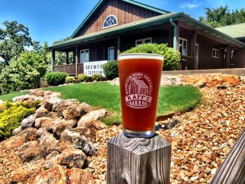 A glass of amber beer sits on a wooden post, with a brewery building and landscaped rocks in the background.