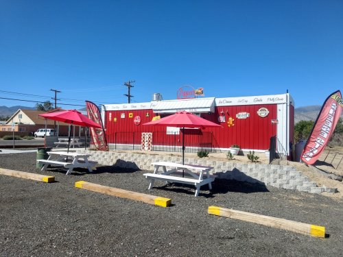 A red food stand with picnic tables and umbrellas, surrounded by gravel and mountains under a clear blue sky.