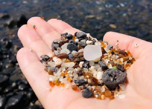 A hand holding a variety of small pebbles and colorful sand by the water's edge.