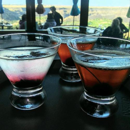 Three cocktails on a tray, one with a cherry, set against a scenic background of hills and people in a restaurant.