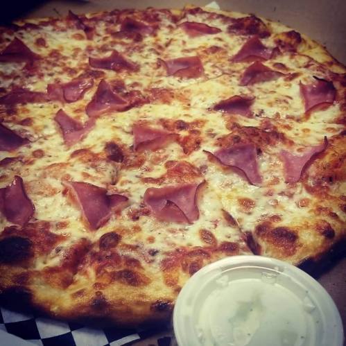 A close-up of a pizza topped with cheese and slices of ham, accompanied by a small container of dipping sauce.