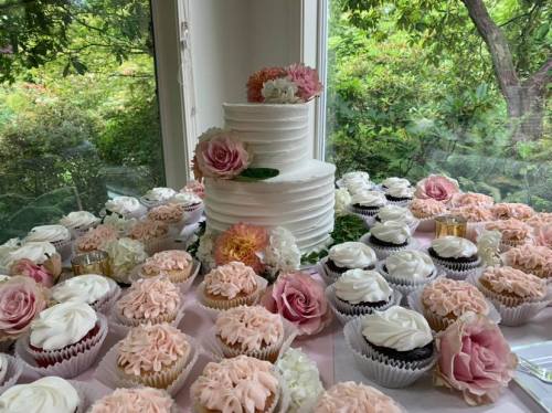 A tiered white cake surrounded by an assortment of decorated cupcakes and fresh flowers on a table.