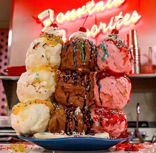 A towering ice cream sundae with various flavors, topped with sprinkles, cherries, and colorful syrup, against a neon sign.