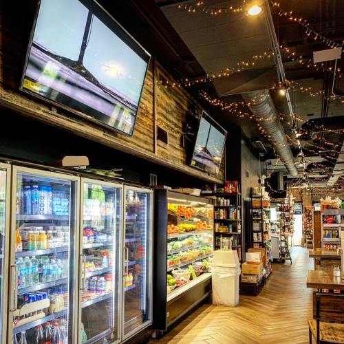 Interior of a grocery store featuring shelves of products, coolers with drinks, and TVs displaying content.