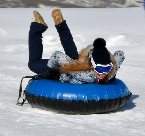 A person in winter gear joyfully sledding down a snowy hill on a blue inflatable tube.