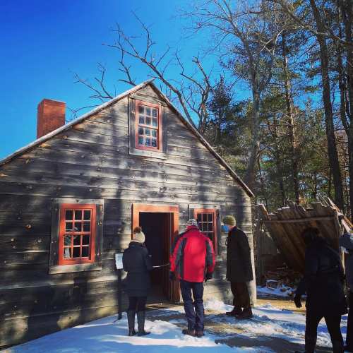 Visitors approach a rustic wooden cabin surrounded by snow and trees on a clear winter day.