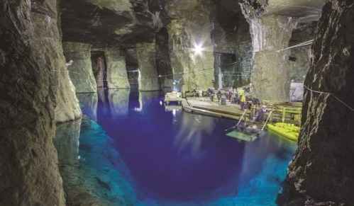 A cavernous underground space with deep blue water, illuminated by lights, and people working on a wooden platform.
