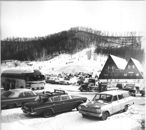 A snowy landscape with skiers on a hill, vintage cars parked in front of a chalet-style building.