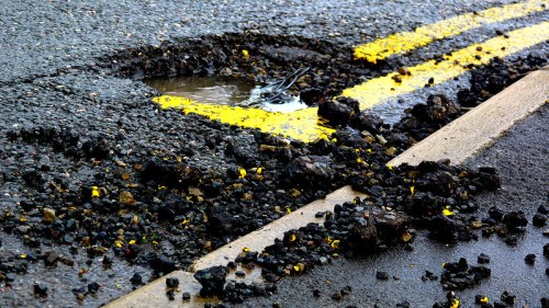 A pothole on a road with yellow lines, surrounded by loose asphalt and a small puddle of water.