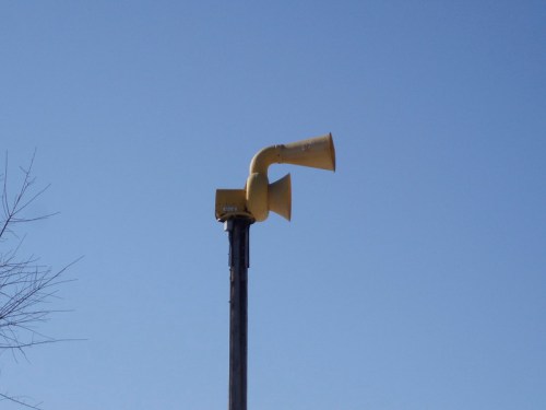 A yellow outdoor warning siren mounted on a tall pole against a clear blue sky.