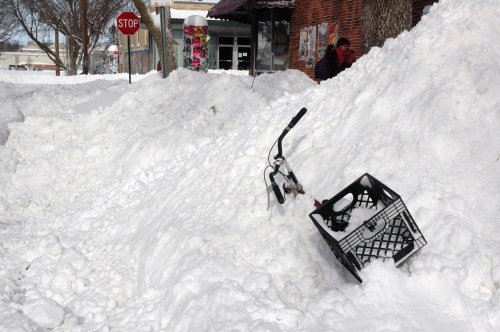 A snowy street scene with a bike partially buried in snow and a plastic crate nearby, with a stop sign in the background.