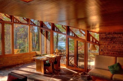 Cozy interior with large windows overlooking a forested area and lake, featuring a wooden table and comfortable seating.