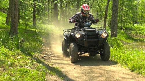 A person riding an ATV on a dirt trail surrounded by trees, kicking up dust in a sunny forest setting.