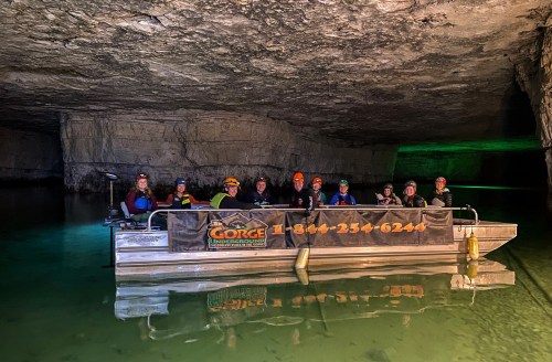 A group of people in helmets on a boat inside a dark underground cave with green lighting and calm water.