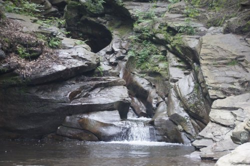 A serene rocky landscape with a small waterfall flowing into a calm pool of water surrounded by lush greenery.