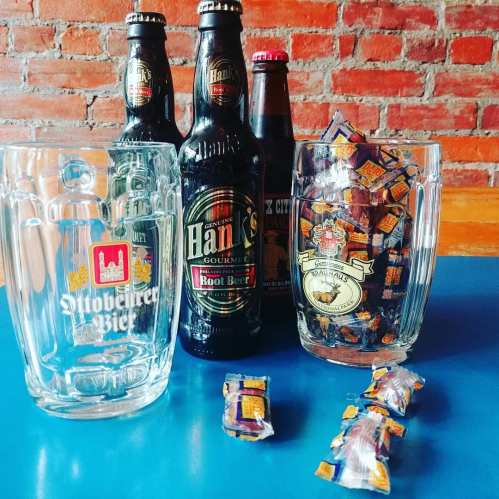 A table with root beer bottles, a glass mug, and a jar of candy, set against a brick wall background.