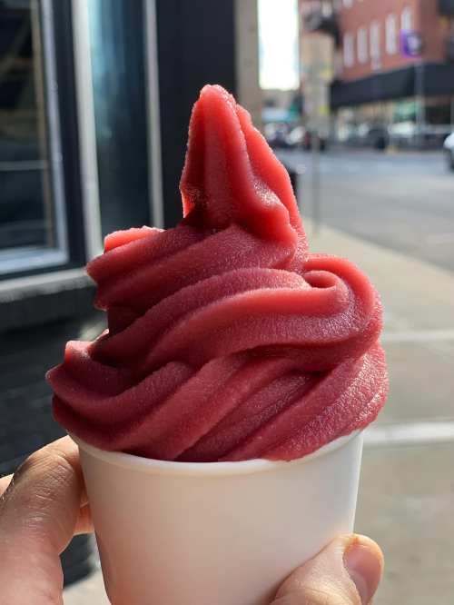 A hand holding a cup of vibrant red sorbet with a soft-serve swirl, set against a city street backdrop.