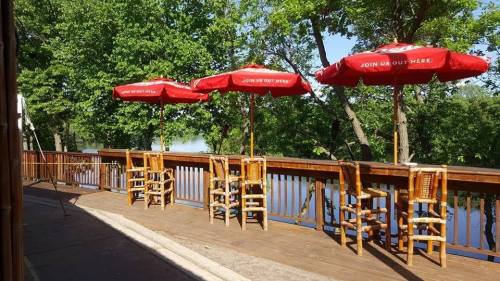 Outdoor seating area with wooden chairs and red umbrellas overlooking a serene river surrounded by trees.
