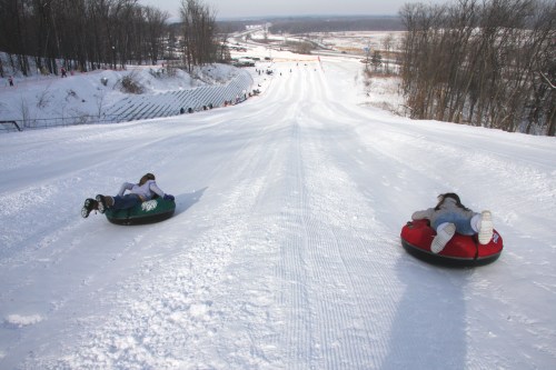Two people slide down a snowy hill on inflatable tubes, surrounded by trees and a distant view of a road.
