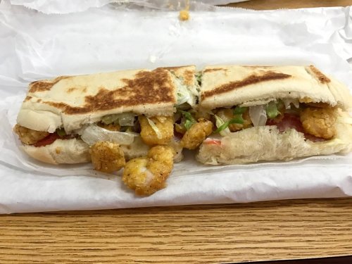 A toasted sandwich filled with fried shrimp, lettuce, and tomato, served on a white paper wrapper.