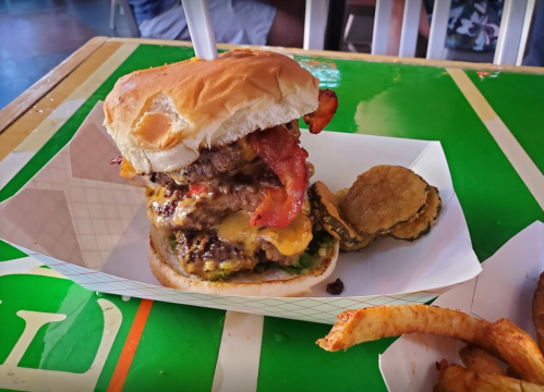 A towering burger with multiple beef patties, melted cheese, bacon, and pickles, served with fries on a colorful table.