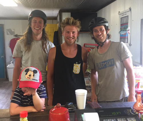 Four young men, two wearing helmets, smile at the camera in a casual indoor setting. A child hides their face.