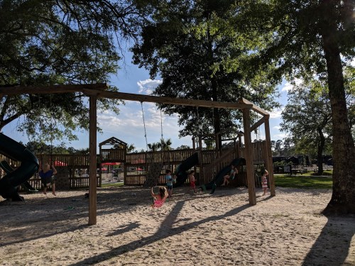 Children playing on swings and slides in a sunny playground surrounded by trees.