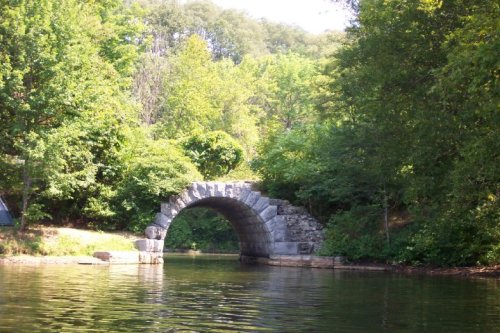 A stone arch bridge over calm water, surrounded by lush green trees and foliage.
