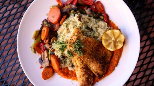 A plate of seasoned fish fillets on a bed of risotto, surrounded by colorful sautéed vegetables and a lemon wedge.