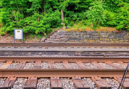 View of train tracks with gravel, surrounded by lush green foliage and a stone wall in the background.