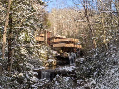 A modern wooden house nestled in a snowy forest, overlooking a small waterfall and surrounded by trees.