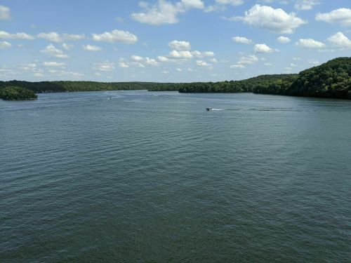 A serene river scene with lush green banks and a boat gliding across the water under a blue sky with fluffy clouds.